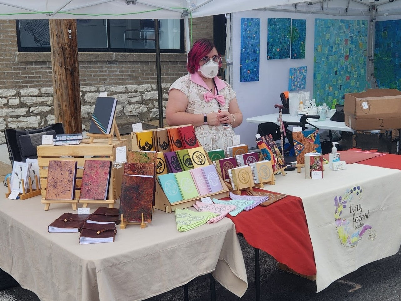 Michio, a light skinned mixed Asian American with bright magenta hair, stands behind their table at an art fair wearing an N95 and a handmade dress. On their table are a variety of handmade books and furoshiki.