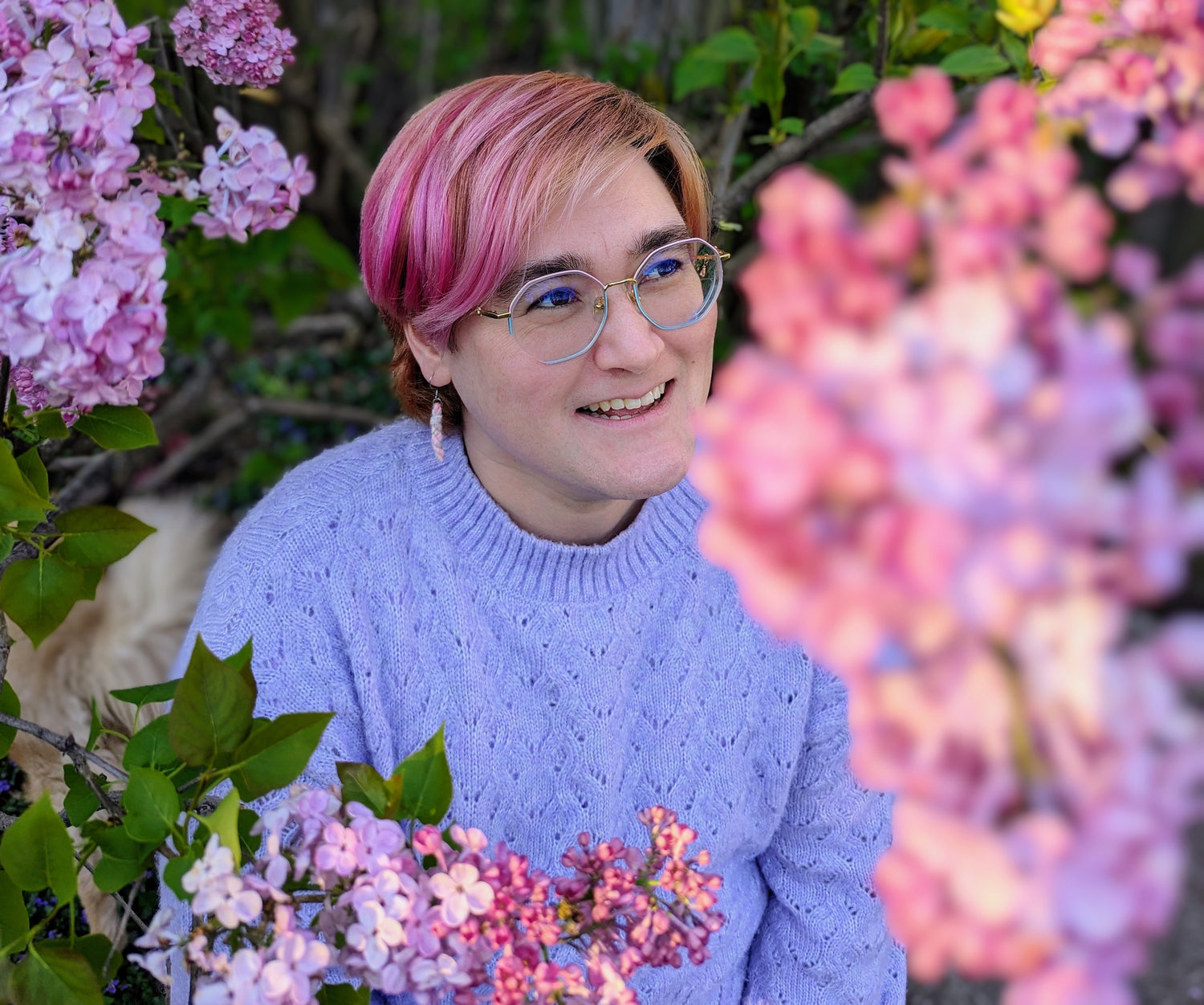 Michio, a mixed Asian American sits smiling surrounded by lilac flowers. They have a pale complex, glasses, and short pink hair. They are wearing a pastel purple knit sweater with mini yarn skein earrings.