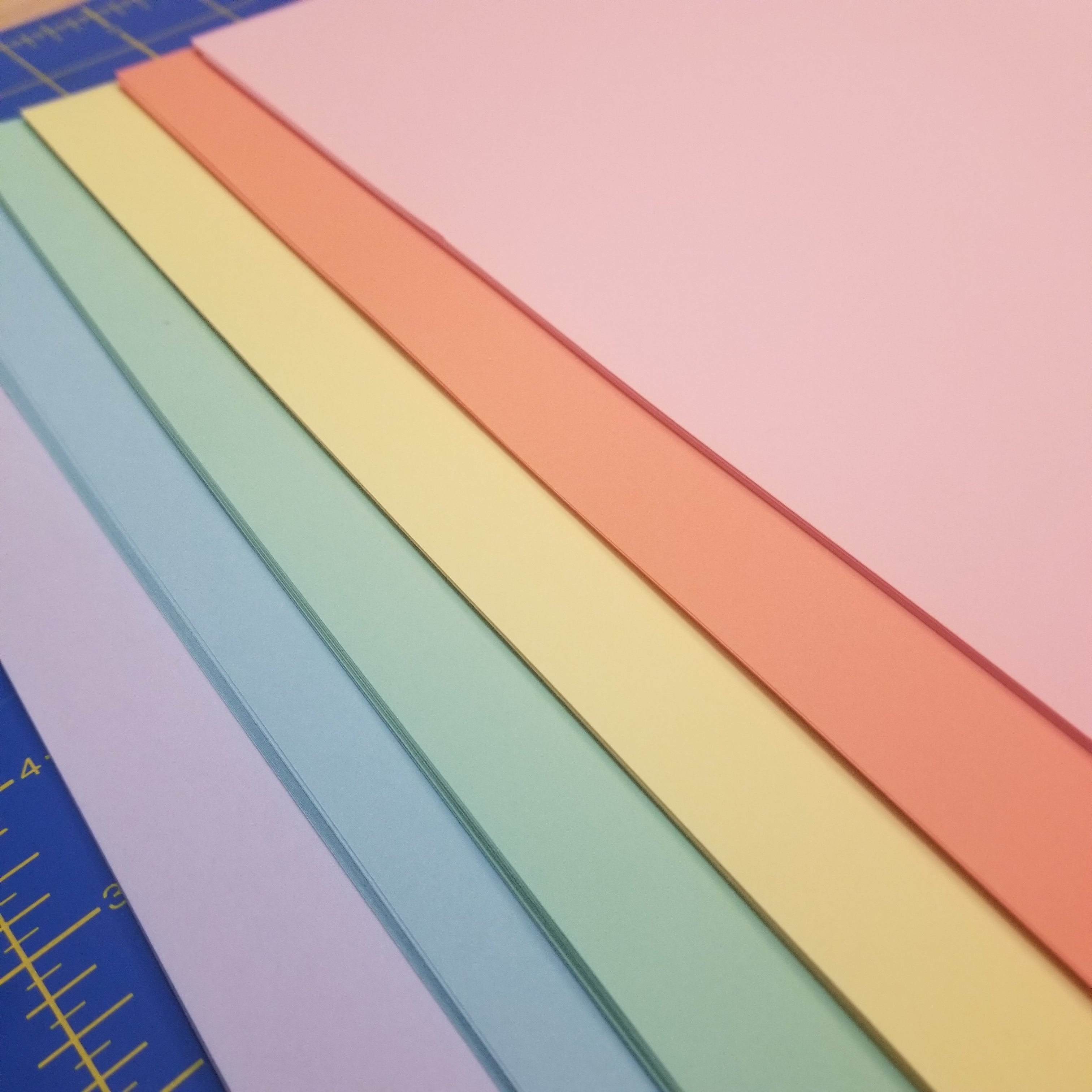 A range of pastel rainbow color papers overlapping each other on a blue cutting mat, seen in the top left and right corner of the photo.