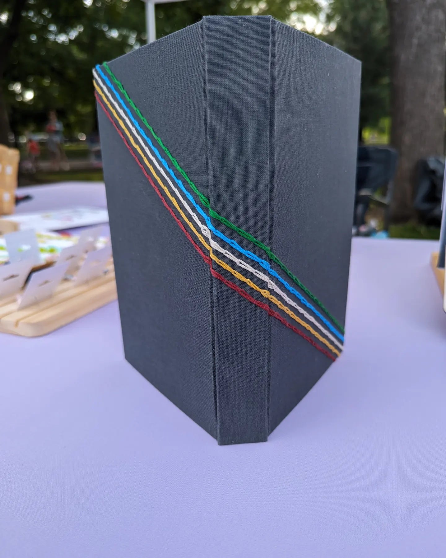 Hardbound blank journal standing open with the spine and covers facing the camera. The book is covered in dark grey fabric with five lines of chain stitches, in red, gold, white, blue, and green. The stitches go from the top left corner of the back cover, crossing over the spine, and ending at the right bottom corner of the front cover creating the Disability Pride flag. 