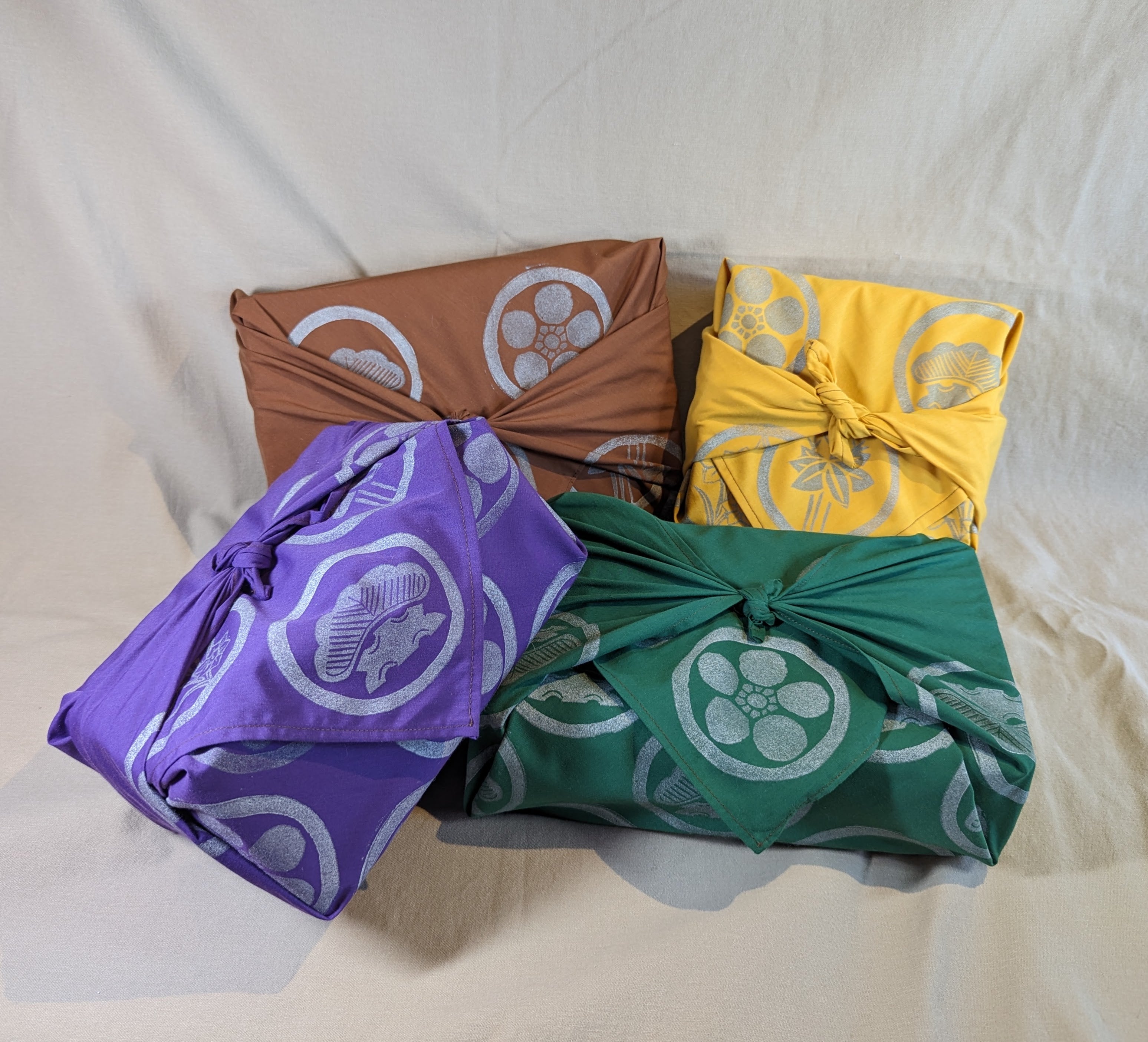 Four rectangular packages are wrapped in furoshiki, Japanese wrapping cloths. Each box has a different color cloth. The clothes are brown, yellow, purple, and green with a variety of crests on each one.