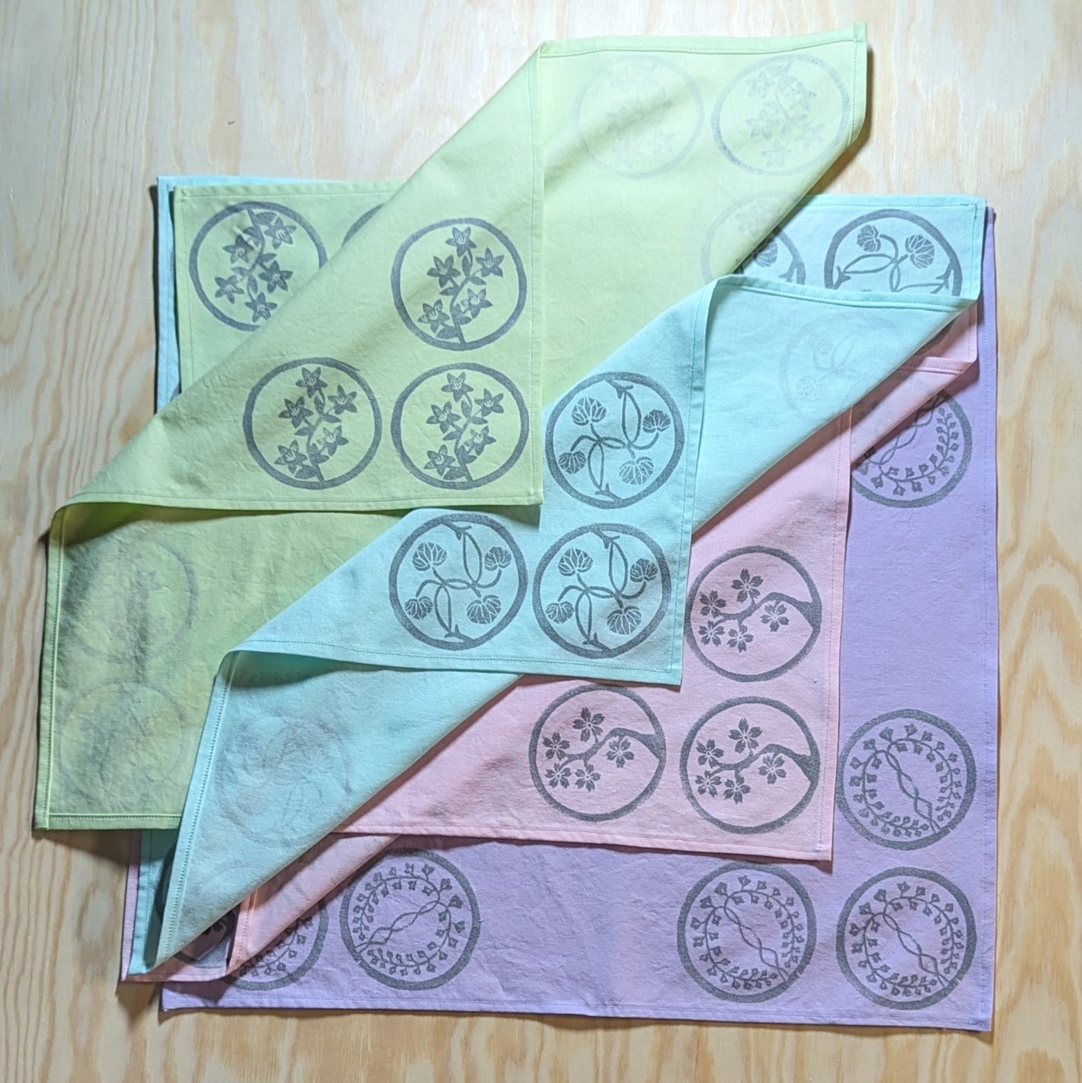 Four pastel wrapping cloths with silver linocut prints stylized to look like Japanese crests are folded overlapping one another. Each wrapping cloth is showing off the print on the bottom right corner which consists of three crests forming a triangle. From top to bottom: green with azaleas, blue with hollyhock leaves, pink with cherry blossoms, and purple with wisteria.