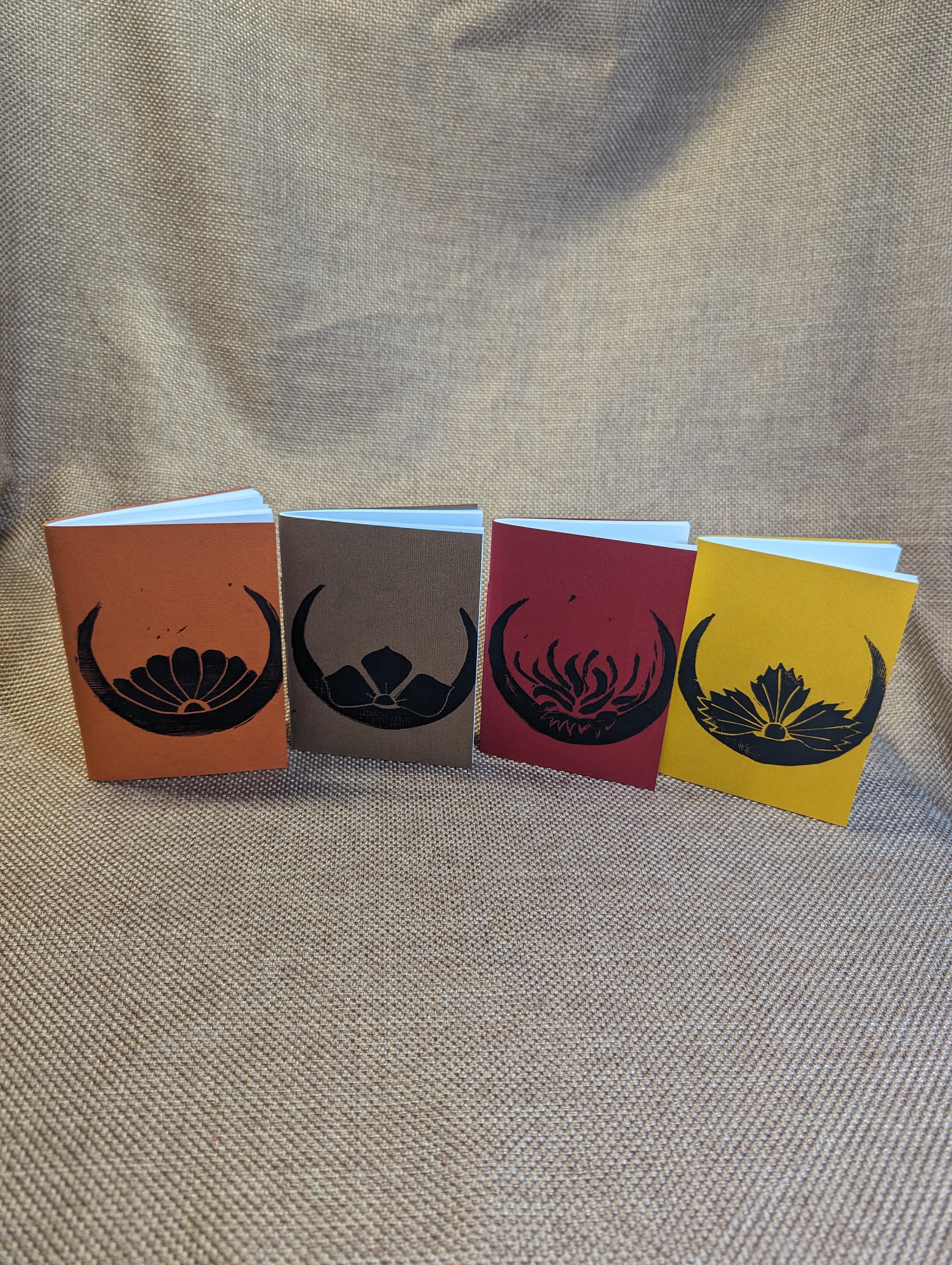 Four pamphlets with black linocut prints stylized to look like Japanese crests sitting on  a crescent, from left to right: orange with chrysanthemum, light brown with Chinese bellflower, red with spider lily, and yellow with dianthus.