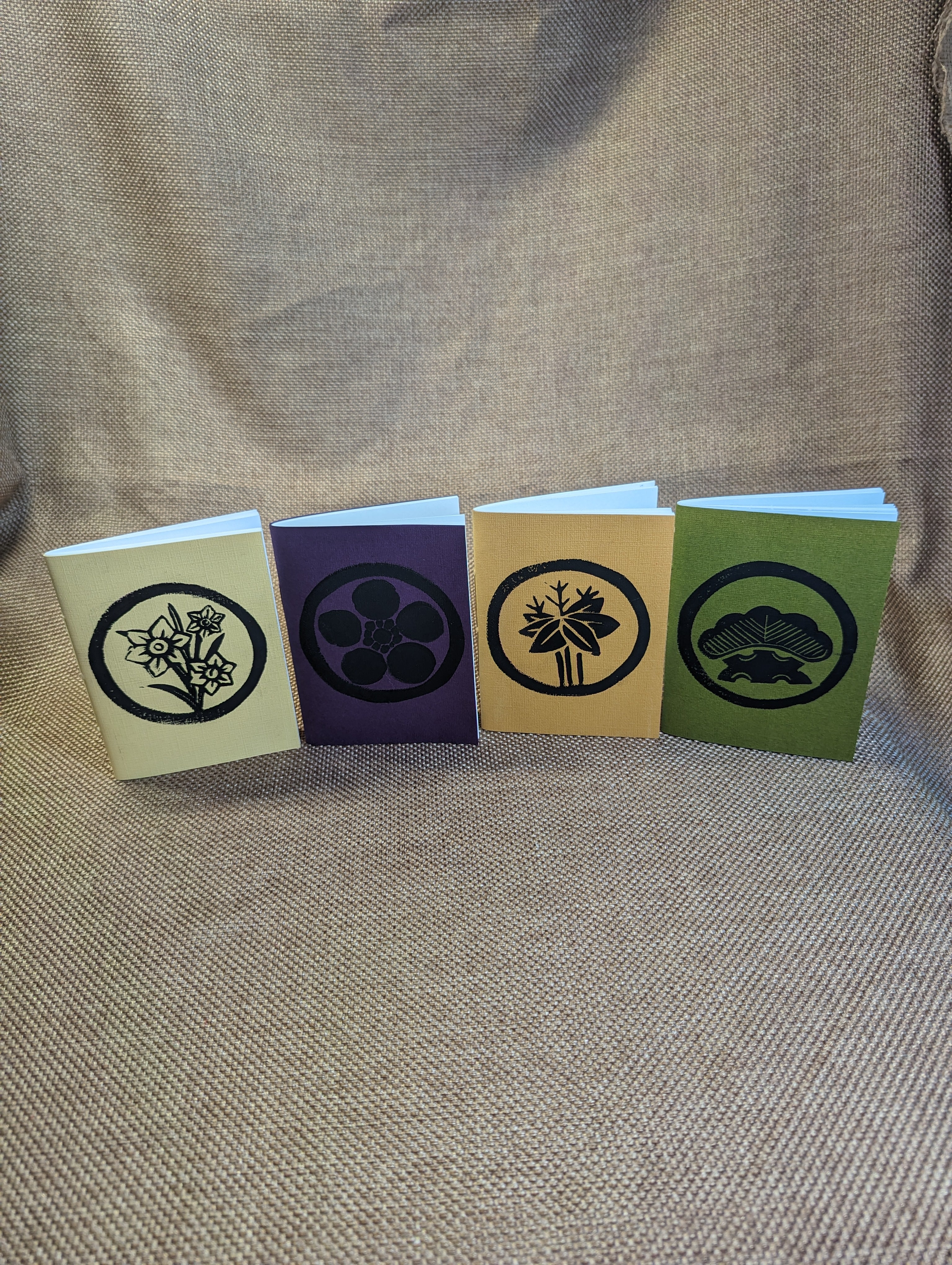 Four pamphlets with black linocut prints stylized to look like Japanese crests centered in a circle, from left to right: pale yellow with daffodil, dark purple with plum, wheat yellow with bamboo, and green with pine.