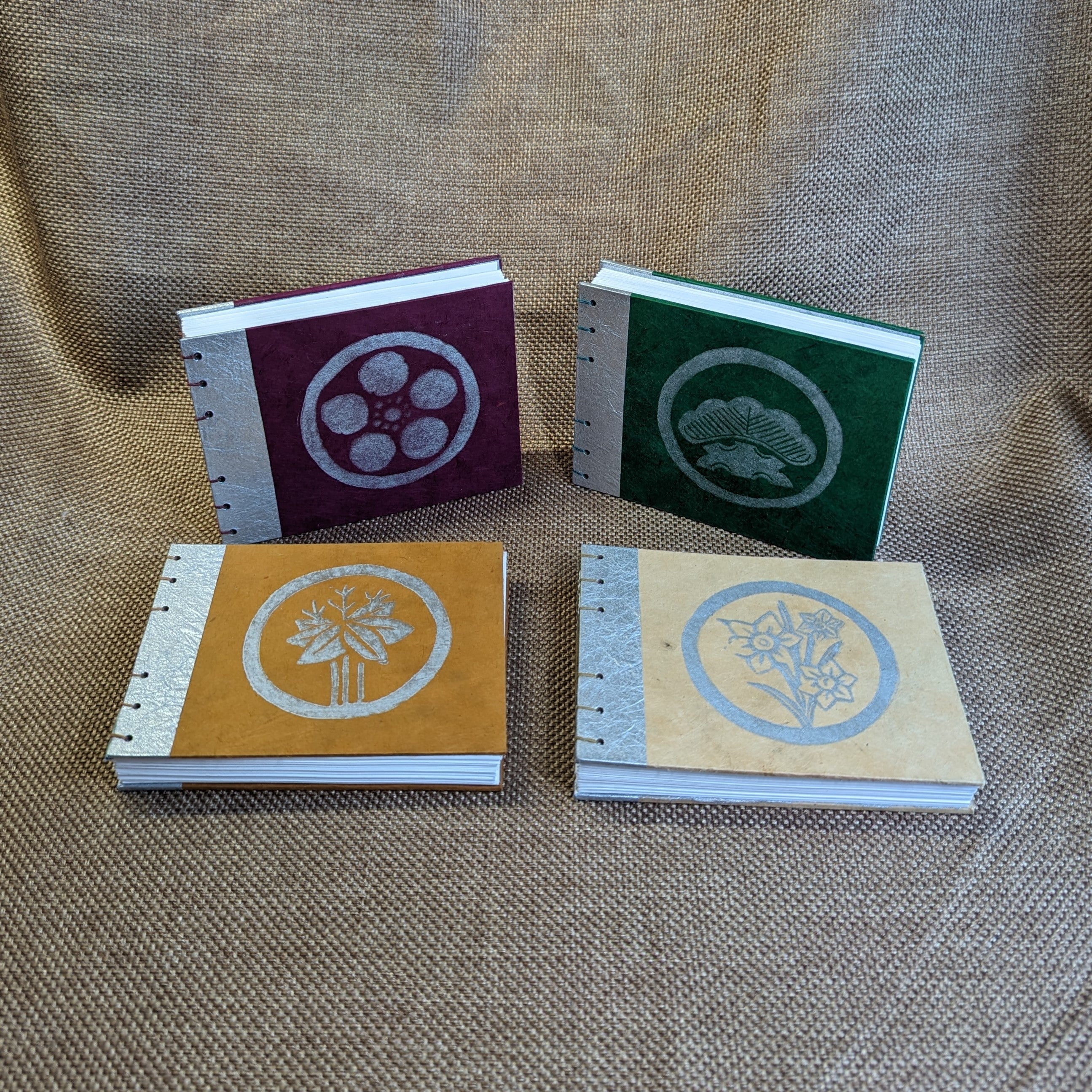 Four exposed binding journals with metallic silver paper on the spine side. Each has a silver linocut print stylized to look like Japanese crests centered in a circle, from left to right: pale yellow with daffodil, dark purple with plum, wheat yellow with bamboo, and green with pine.