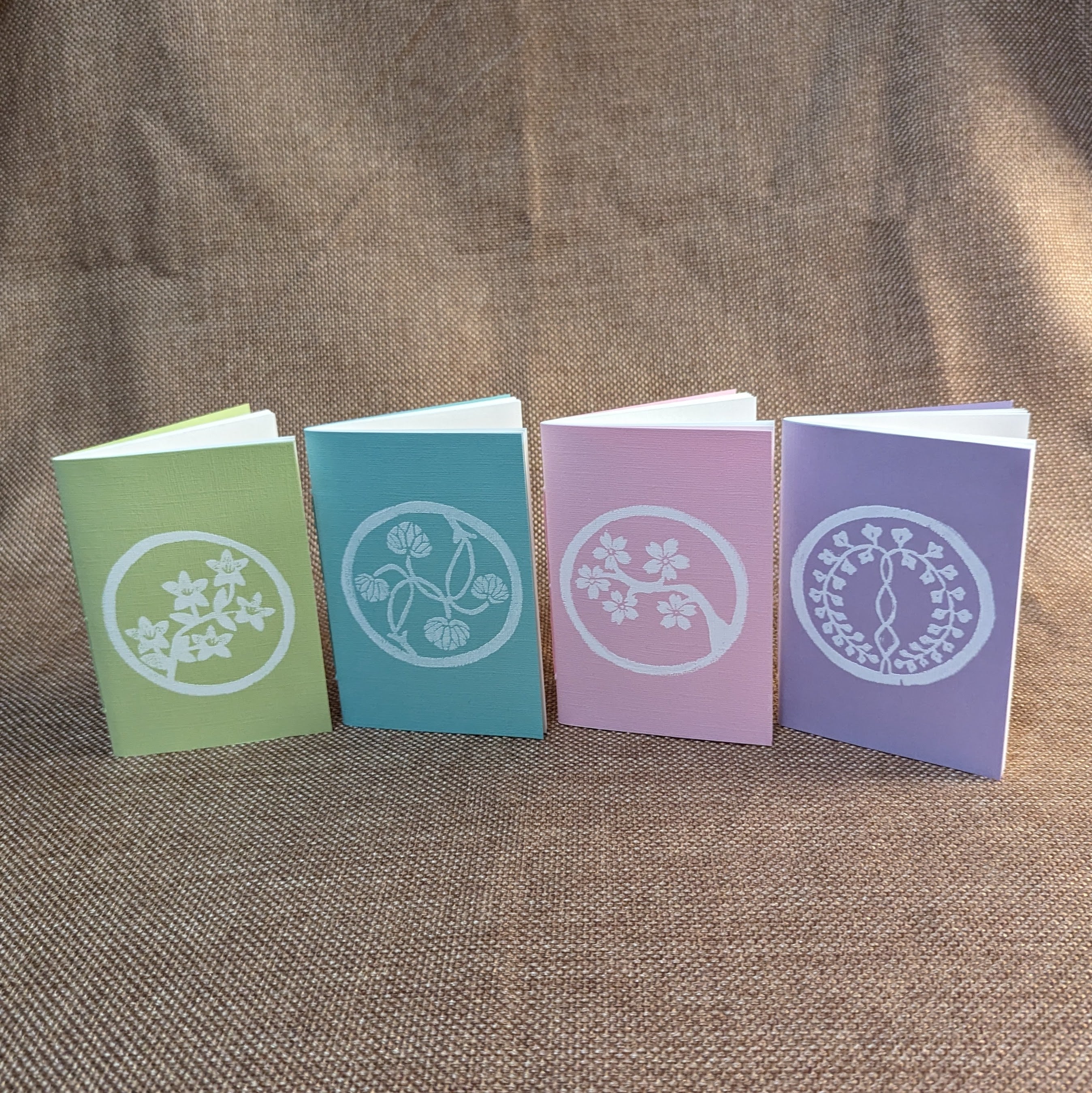 Four pastel pamphlets with white linocut prints stylized to look like Japanese crests, from left to right: green with azaleas, blue with hollyhock leaves, pink with cherry blossoms, and purple with wisteria.